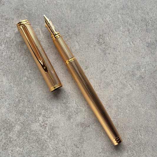 A gold-colored fountain pen with a textured grip and matching cap, displayed diagonally on a gray textured surface.