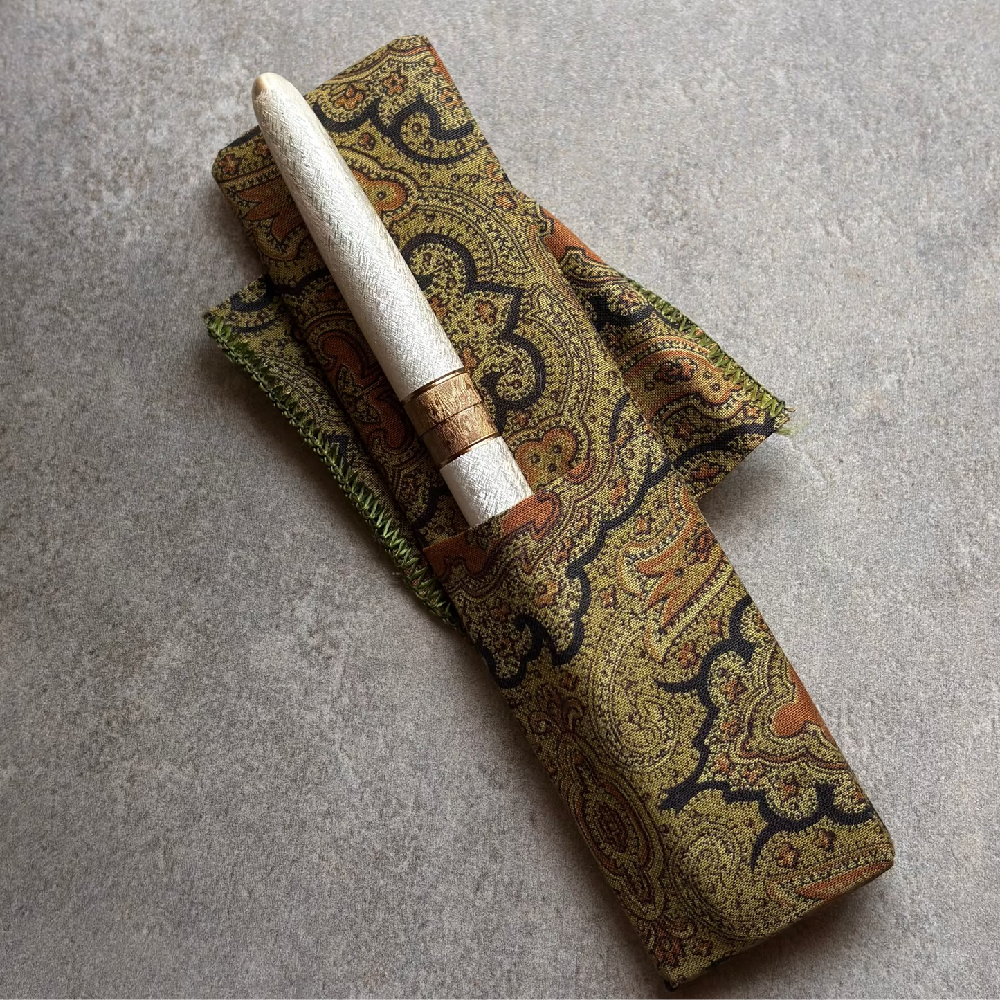 A white fountain pen with a gold colored band, resting on a matching patterned fabric sheath with green trim, placed on a textured gray surface.