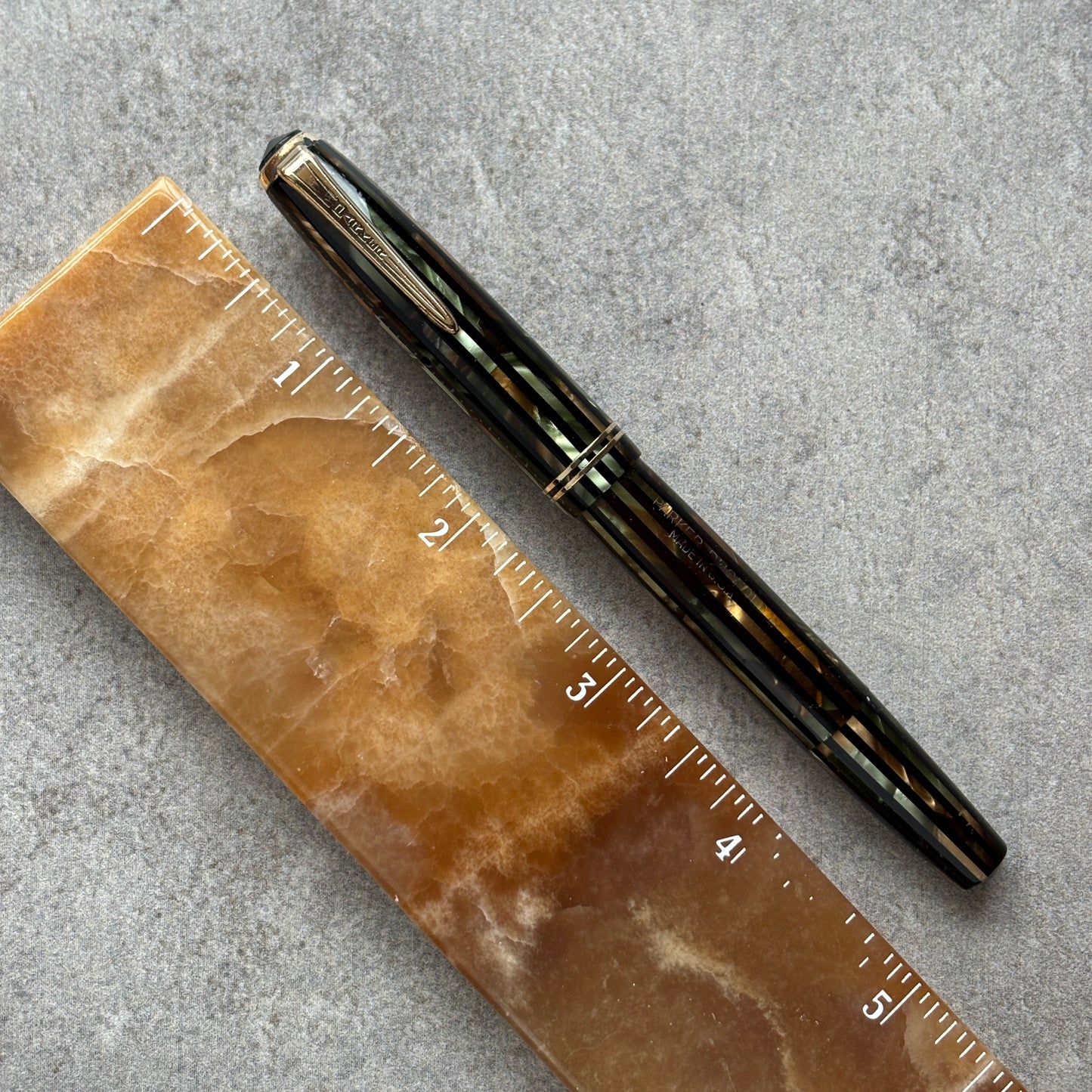 Black and green pen next to a brown marble ruler on a gray surface