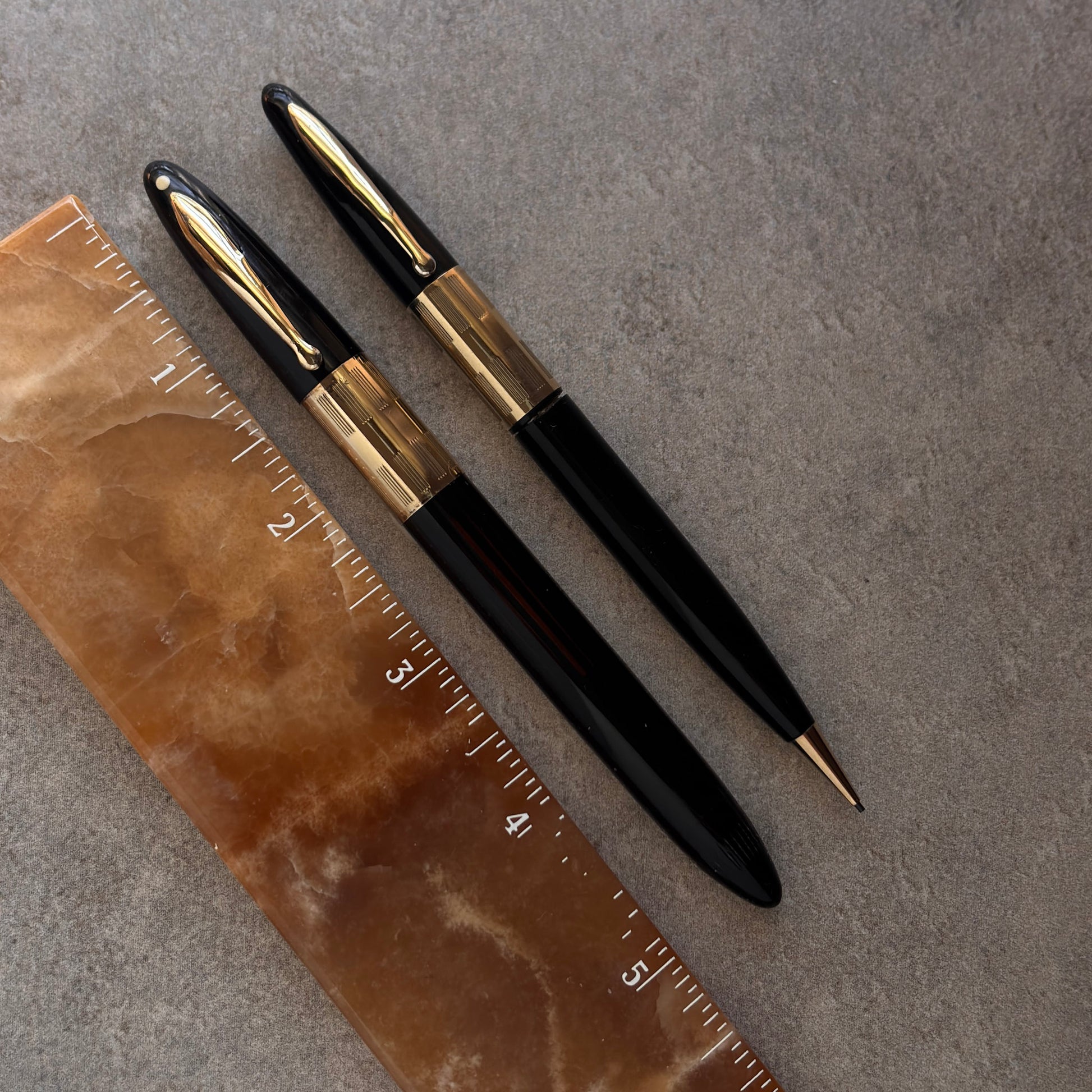 Two black and gold pens on a textured surface with a ruler for scale.