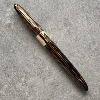 A luxury fountain pen with a dark brown and black striped wood-grain body and polished gold-tone metal accents, resting diagonally on a textured gray concrete surface.