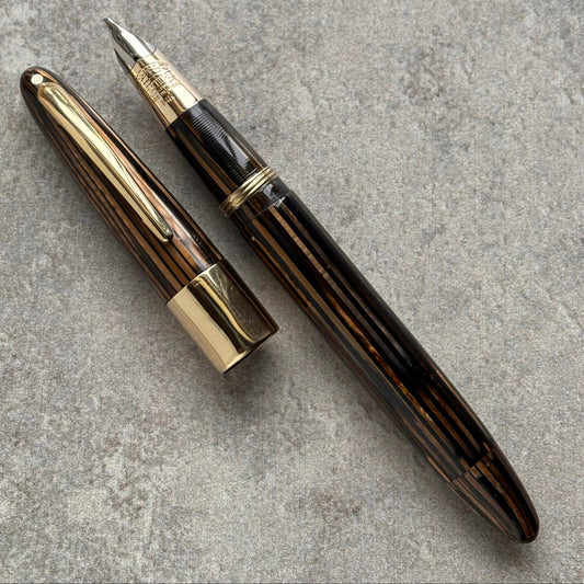 A luxury pen with a dark brown and black striped wood grain body and polished gold-tone metal accents, shown with its cap off on a textured gray background.