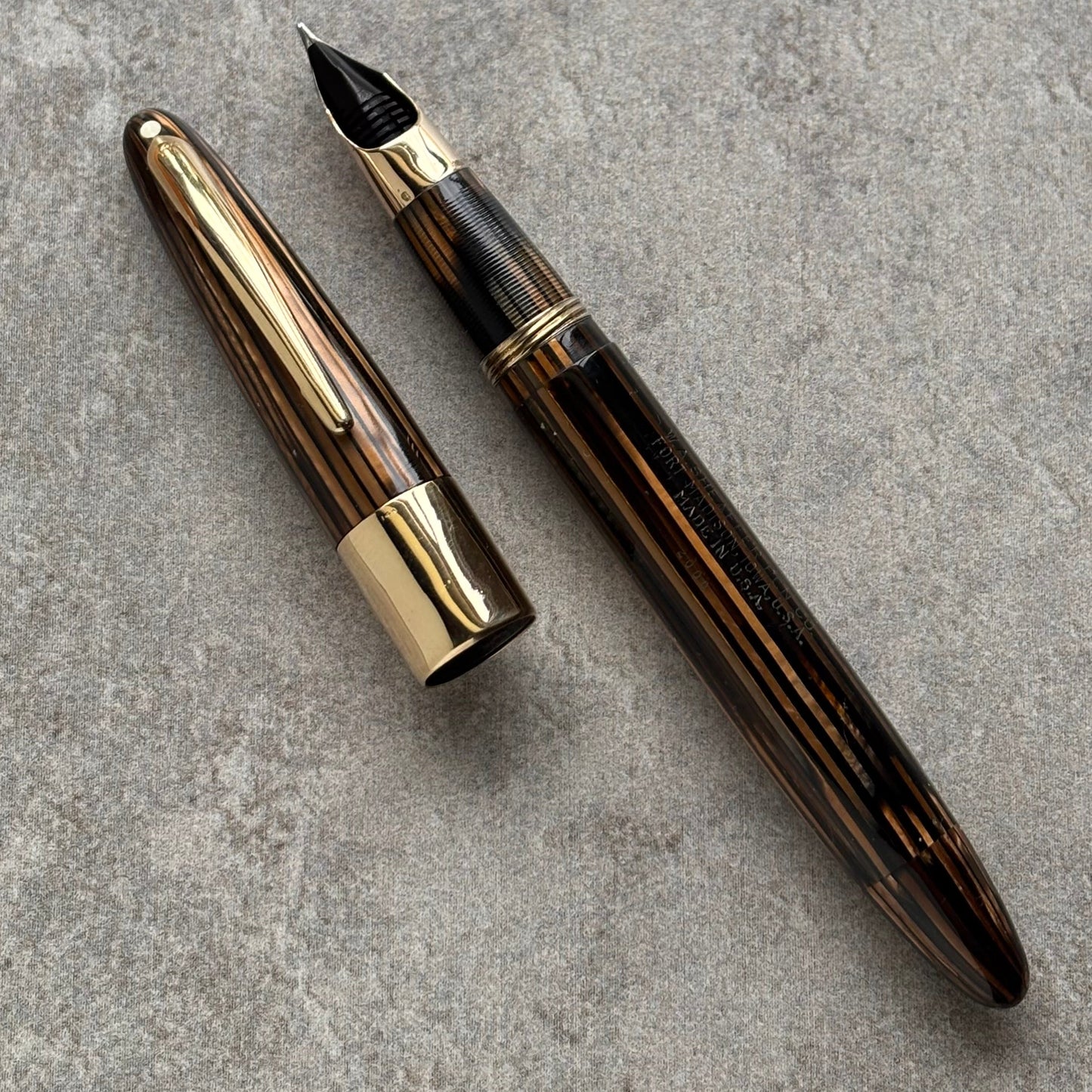 A luxury fountain pen with a dark brown wood-grain patterned barrel and polished gold-tone metal accents, shown with its cap removed and placed beside it on a textured gray concrete surface.