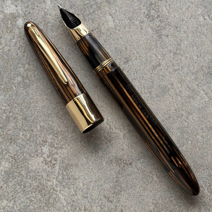 A luxury fountain pen with a dark brown wood-grain patterned barrel and polished gold-tone metal accents, shown with its cap removed and placed beside it on a textured gray concrete surface.
