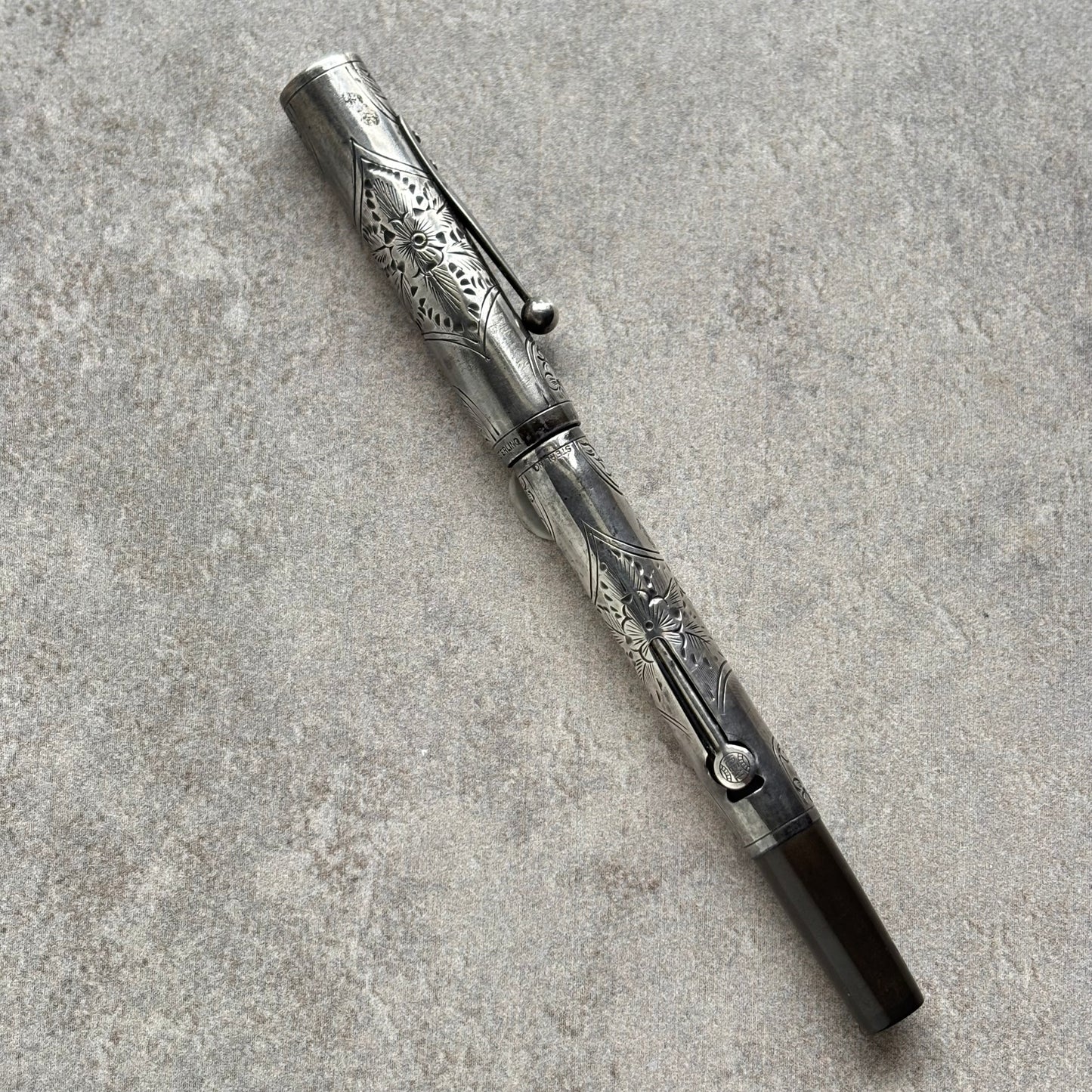 An ornate silver-toned metal pen with intricate floral engravings, resting diagonally on a textured gray stone surface.