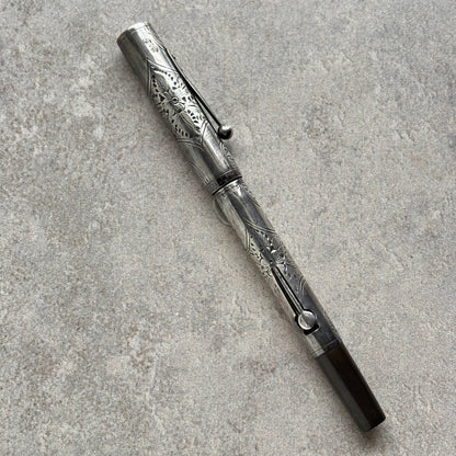 An ornate silver-toned metal pen with intricate floral engravings, resting diagonally on a textured gray stone surface.