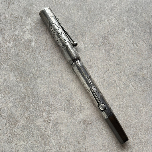 An ornate silver-toned metal pen with intricate floral engravings, resting diagonally on a textured gray stone surface.