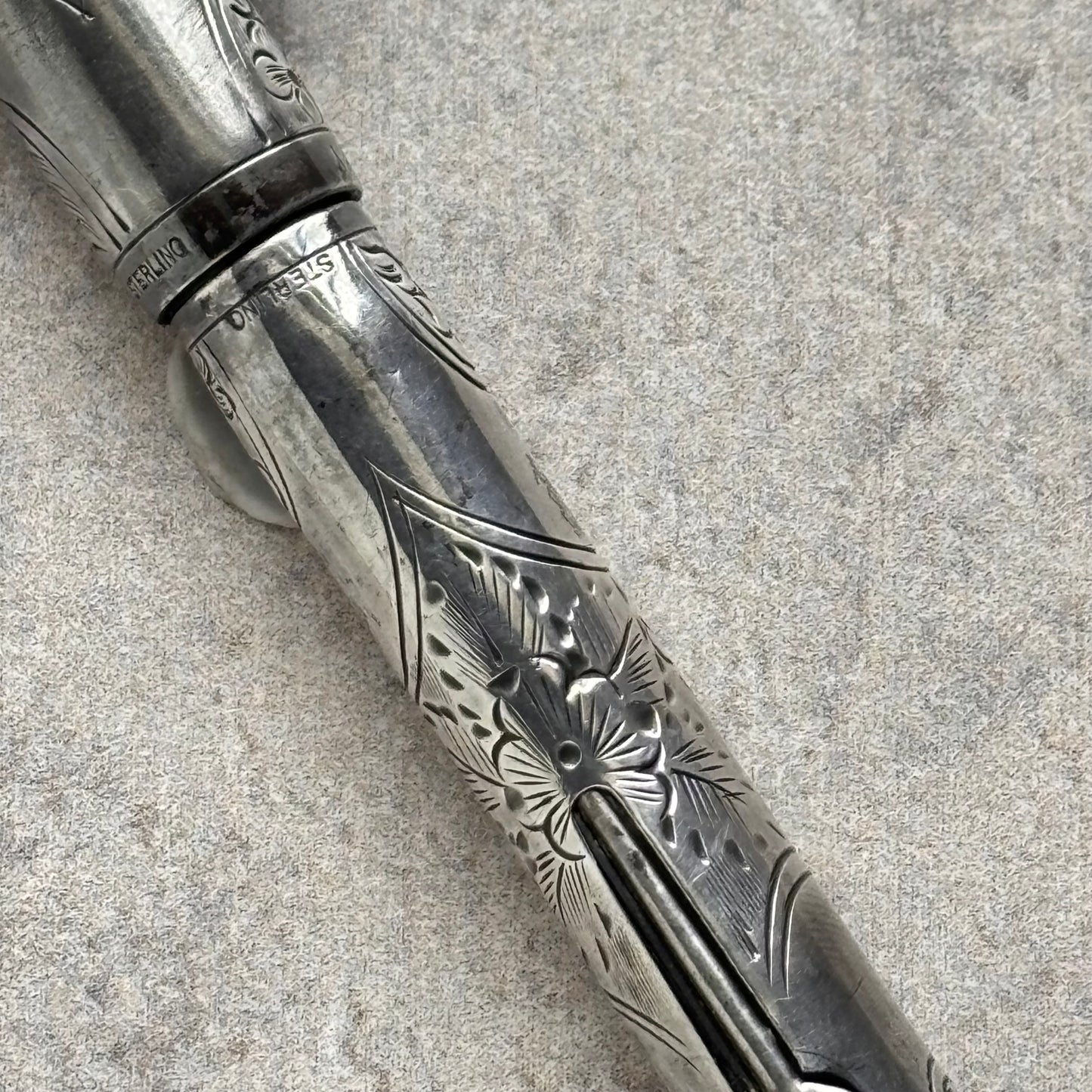 A close-up of an ornate silver-toned metal pen with engraved floral and geometric patterns, resting diagonally on a textured gray surface.