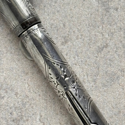 A close-up of an ornate silver-toned metal pen with engraved floral and geometric patterns, resting diagonally on a textured gray surface.