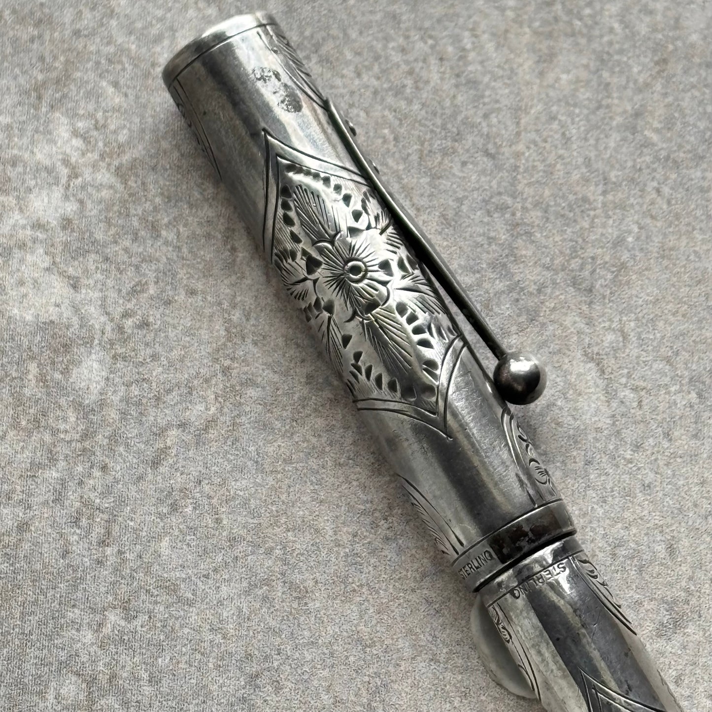 An ornate silver-toned metal pen with intricate floral engravings and a matching cap, photographed diagonally on a textured gray surface.