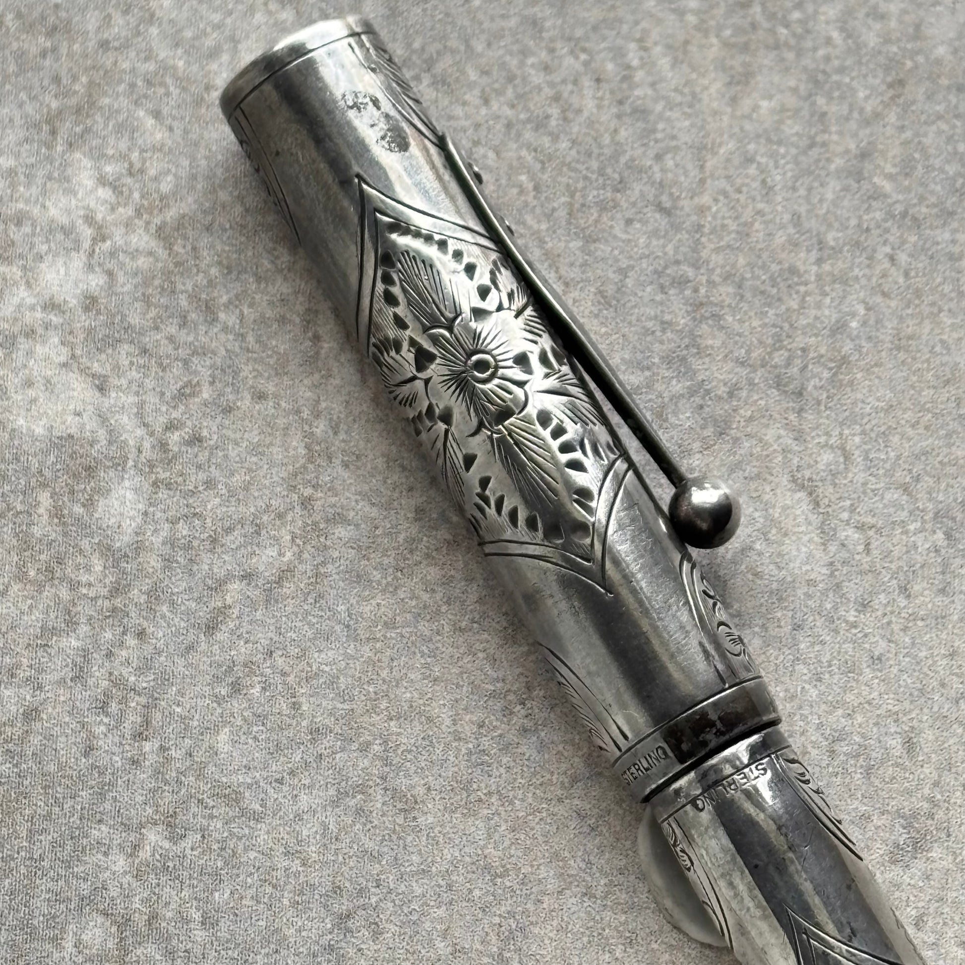 An ornate silver-toned metal pen with intricate floral engravings and a matching cap, photographed diagonally on a textured gray surface.