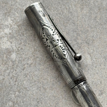 An ornate silver-toned metal pen with intricate floral engravings and a matching cap, photographed diagonally on a textured gray surface.