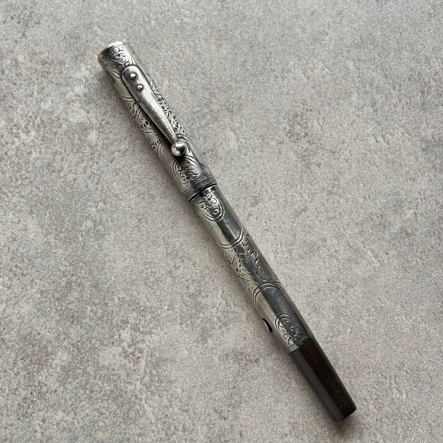 An ornate silver-toned metal pen with detailed floral engravings and a dark brown grip, placed diagonally on a textured gray concrete surface.