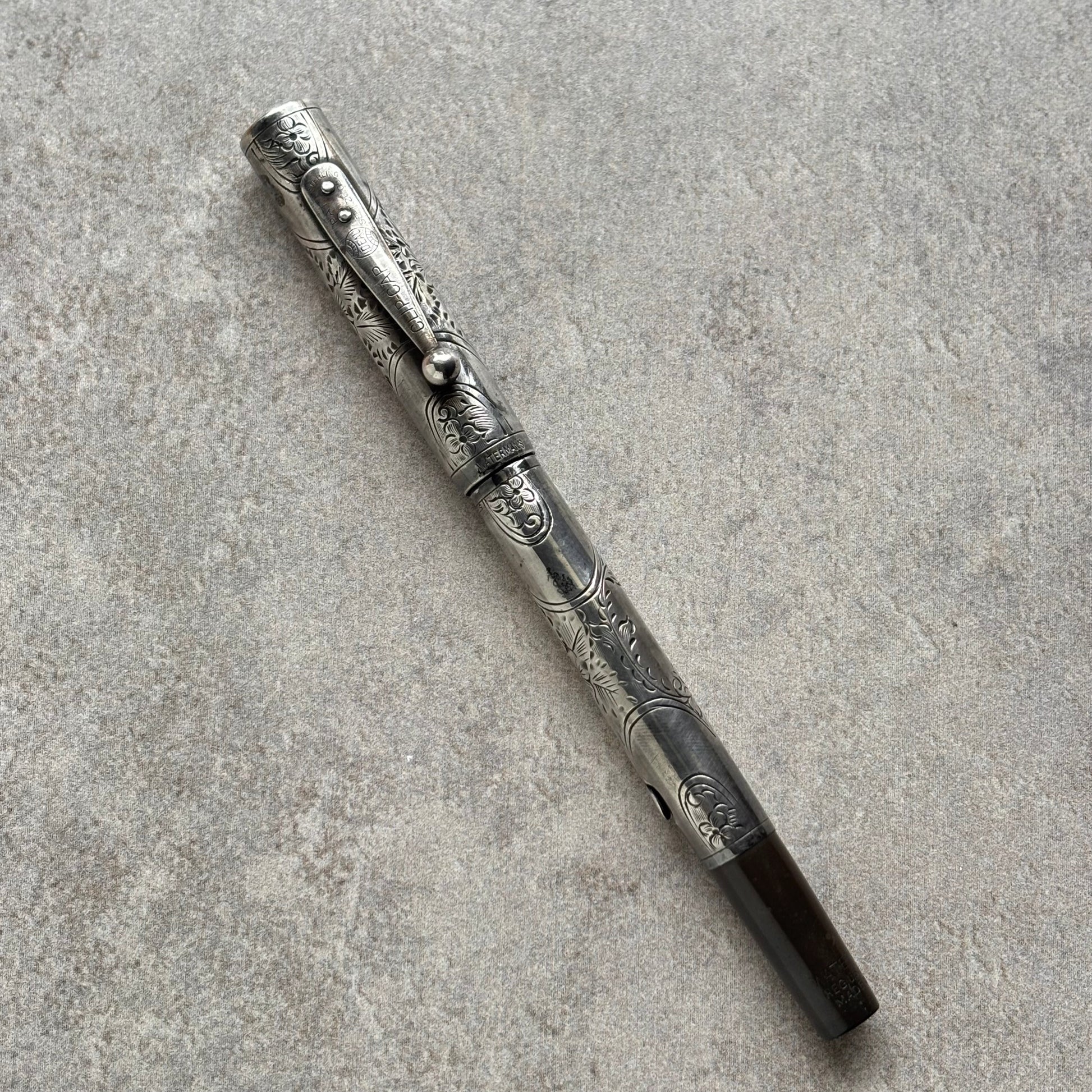 An ornate silver-toned metal pen with detailed floral engravings and a dark brown grip, placed diagonally on a textured gray concrete surface.