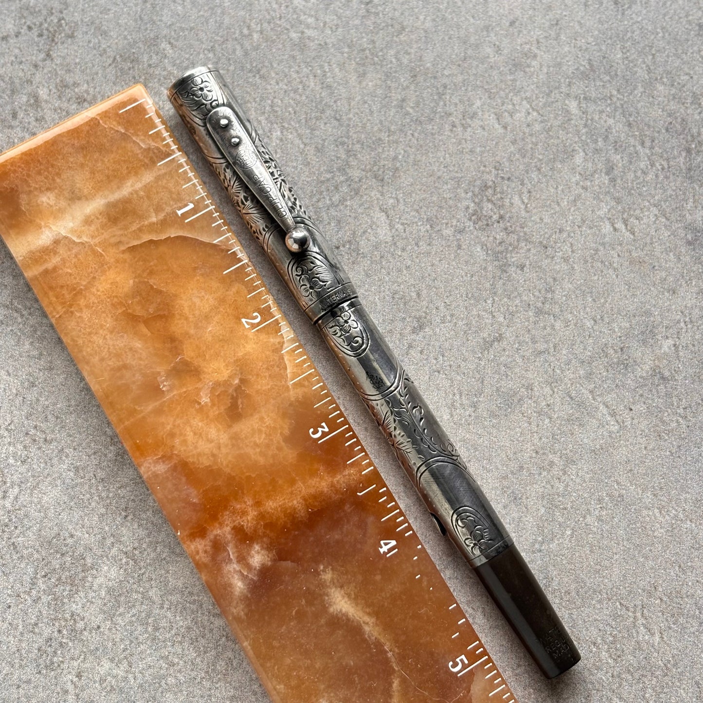 An ornate silver-toned metal pen with intricate engraved patterns and a black grip, shown next to a brown marbled ruler on a gray textured surface.