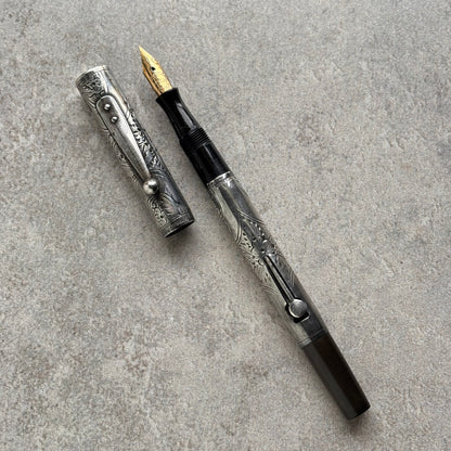 A silver-toned engraved fountain pen with a gold nib and black grip, shown with its cap off on a textured gray surface.