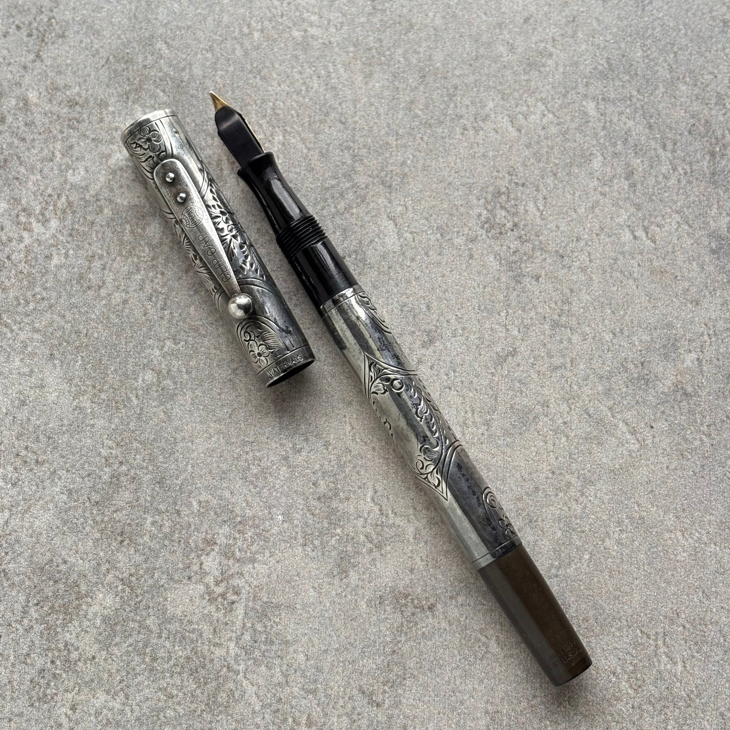 An ornate silver-toned fountain pen with floral engravings, shown with its cap removed and placed beside the barrel on a textured gray surface.
