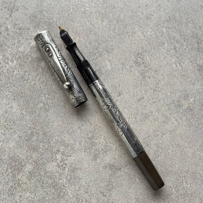 An ornate silver-toned fountain pen with floral engravings, shown with its cap removed and placed beside the barrel on a textured gray surface.