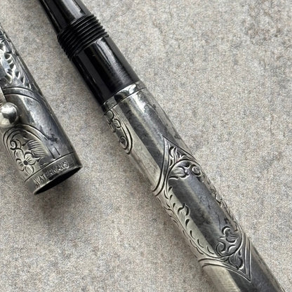A close-up of a silver-toned engraved fountain pen with a black ribbed grip section, shown with its cap beside it on a textured gray surface.