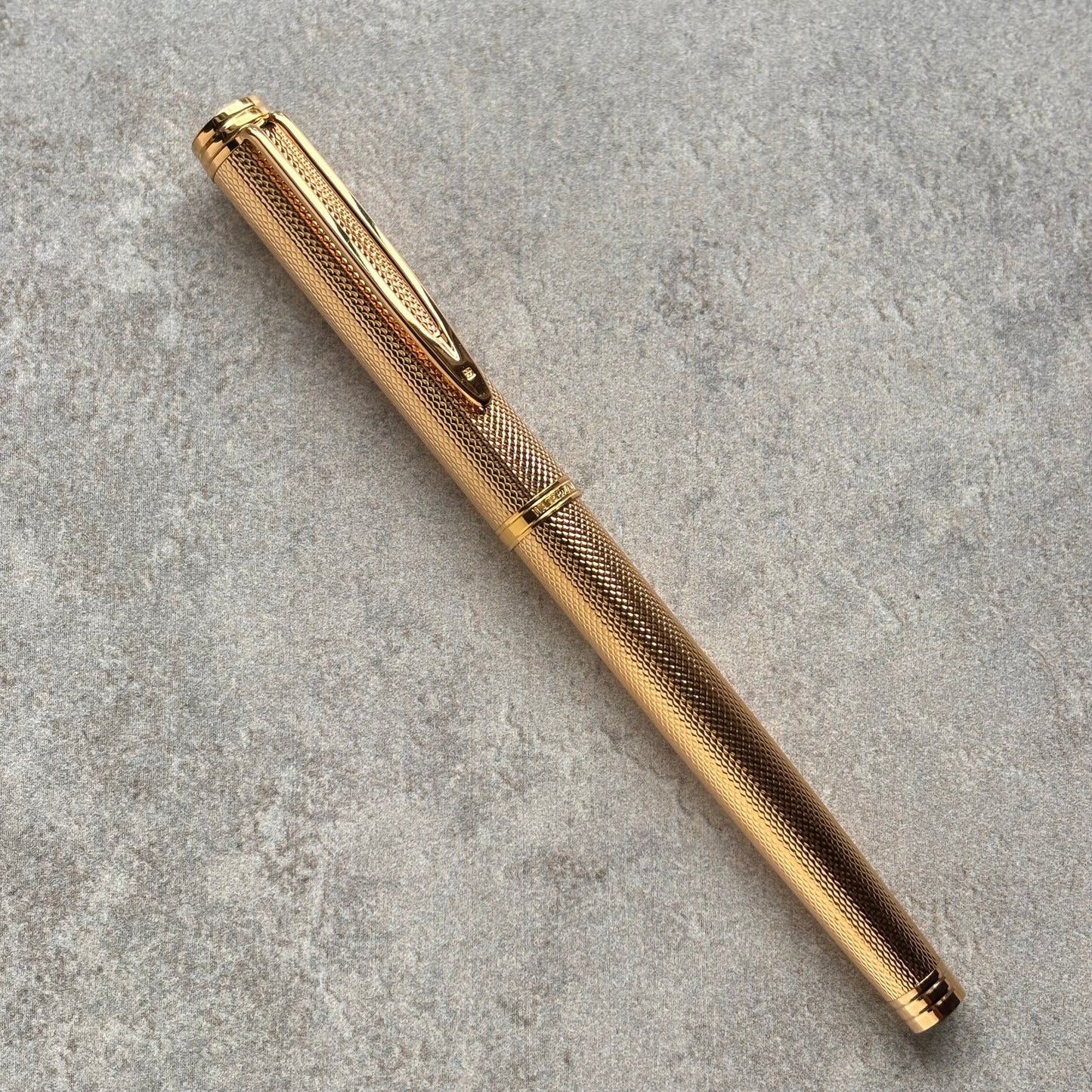 A gold-colored metal pen with a textured grip and a matching gold clip, resting diagonally on a gray textured surface.
