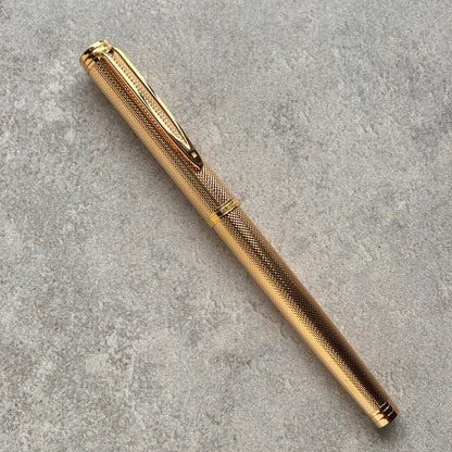 A gold-colored metal pen with a textured grip and a matching gold clip, resting diagonally on a gray textured surface.