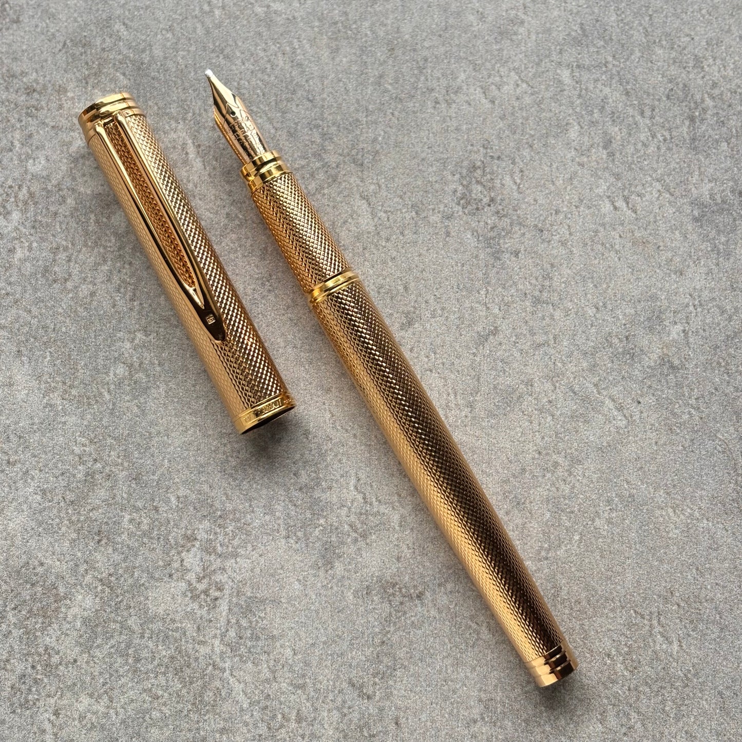 A gold-colored fountain pen with a textured grip and matching cap, displayed diagonally on a gray textured surface.