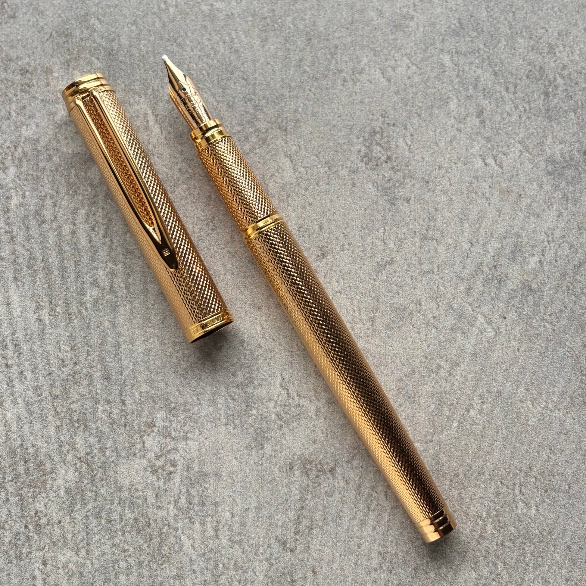 A gold-colored fountain pen with a textured grip and matching cap, displayed diagonally on a gray textured surface.