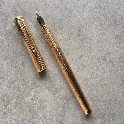 A gold-finished fountain pen with a textured grip and matching cap, shown with the nib exposed on a gray textured surface.