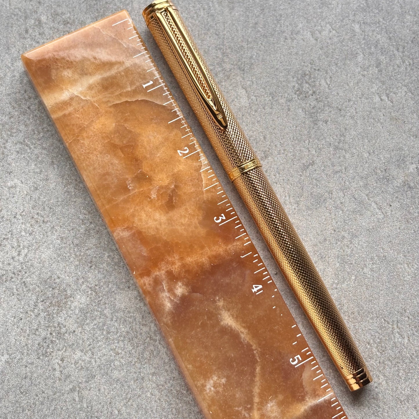 A gold-toned metal pen with a textured grip and clip, placed diagonally next to a translucent amber-brown ruler with white inch markings on a gray stone surface.