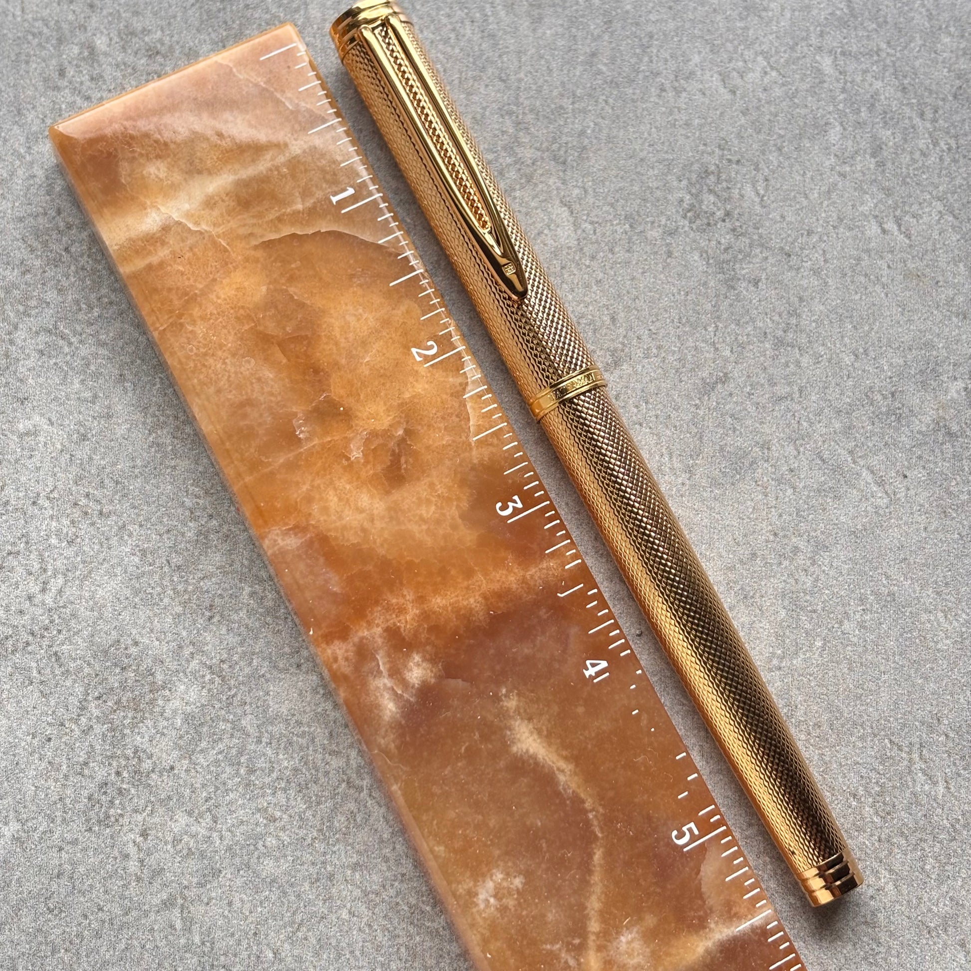 A gold-toned metal pen with a textured grip and clip, placed diagonally next to a translucent amber-brown ruler with white inch markings on a gray stone surface.