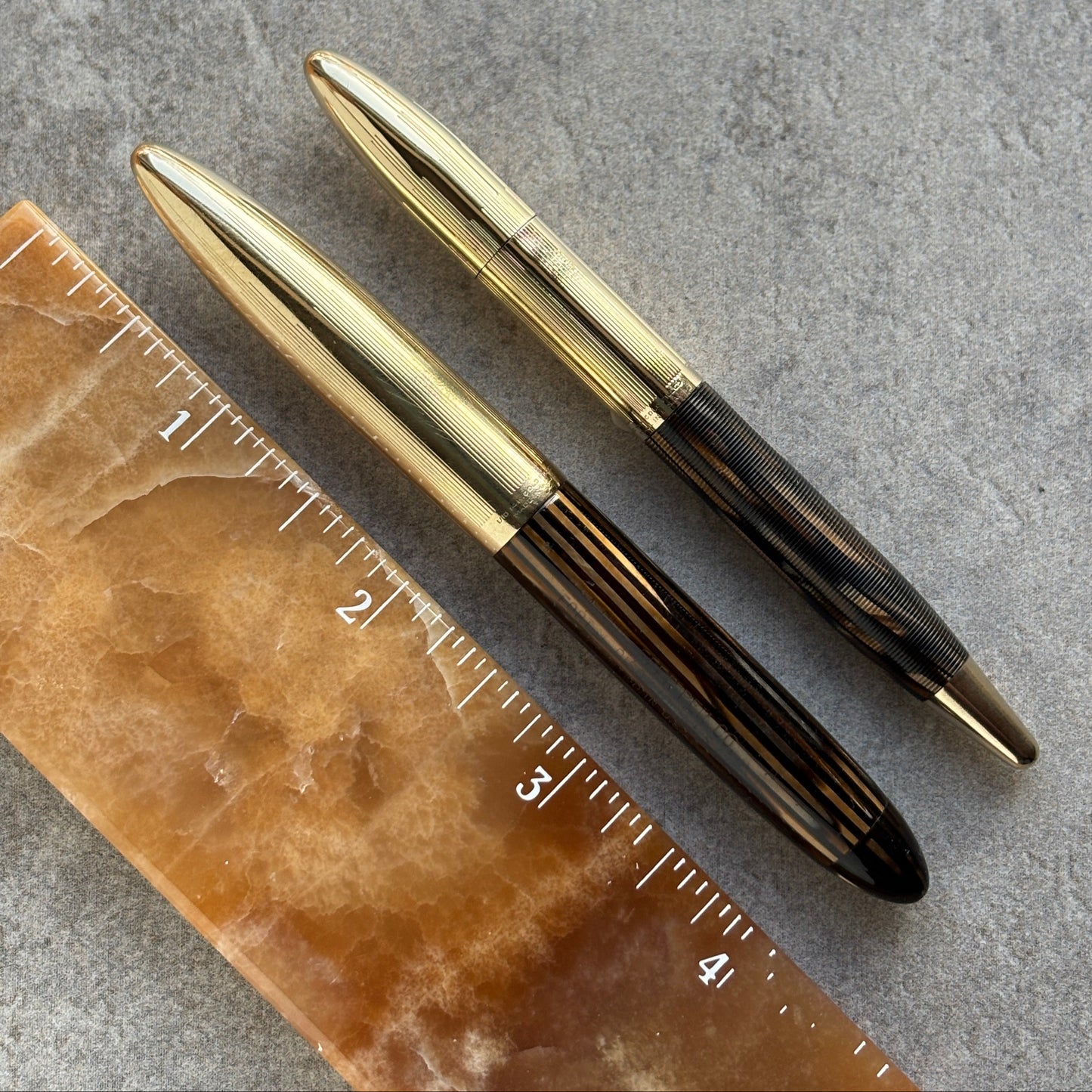 Two gold pens on a textured surface with a ruler for scale.